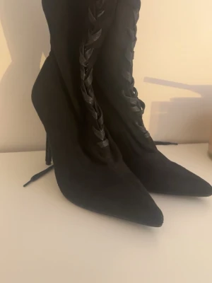 Svarta spetsiga stövletter med snörning - Snygga svarta stövletter med spetsig tå och höga klackar. Skorna har dekorativ snörning framtill med breda band och är tillverkade i ett mockaliknande material. Perfekta för dig som vill ha en edgy och trendig look.