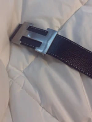 Svart skärp med H-spänne i silver - Stilrent svart bälte i skinn med ett stort H-format spänne i silverfärg. Bältet har vit kontrastsöm längs kanterna och flera hål för justerbar passform. Perfekt accessoar för att lyfta din outfit.