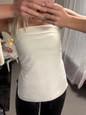 Vit tubtopp med slits från Gina Tricot - Säljer en stilren vit tubtopp från Gina Tricot. Toppen är axelbandslös och har en snygg slits på sidan som ger en trendig look. Materialet är stretchigt och sitter tajt mot kroppen. Perfekt att matcha med jeans eller kjol för en clean outfit.
