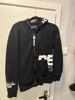 Svart Replay Zip-hoodie med snygga detaljer - Säljer en riktigt fet svart huvtröja med dragkedja från Replay. Den har flera coola detaljer och tryck som gör att den sticker ut från en vanlig basic-hoodie. Storlek XS men passar även S. Hör gärna av dig om du har några frågor eller vill ha exakta mått! 📦 Skickas spårbart