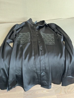 Svart skjorta med broderade fickor - Stilren svart skjorta i silkesliknande material med två stora bröstfickor dekorerade med vitt broderat mönster. Skjortan har lång ärm och knäppning framtill. Perfekt för dig som gillar detaljer och en elegant look.