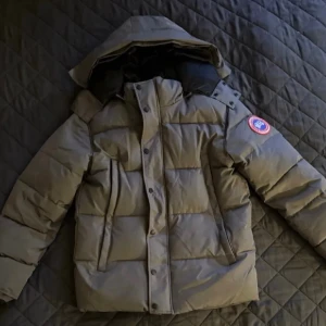 Grå dunjacka från Canada Goose - Snygg grå dunjacka från Canada Goose med huva och klassisk logga på ärmen. Jackan har breda kanaler, hög krage och dragkedja med knappslå. 