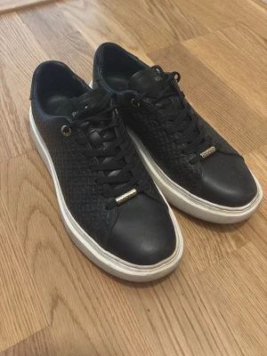 Svarta BOSS sneakers med vit sula - Snygga svarta sneakers från BOSS med präglat mönster på sidorna, gulddetaljer på snörningen och logga i guld på hälen. Skorna har en klassisk låg modell och vit, tjock sula som ger en modern look. Perfekta för dig som gillar stilrena och trendiga sneakers.