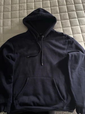 Mörkblå hoodie från H&M - En enkel och stilren mörkblå hoodie från H&M med klassisk känguruficka och justerbar dragsko i huvan. Hoodien har ribbade muddar vid ärmslut och nederkant. Perfekt för en avslappnad och casual look.