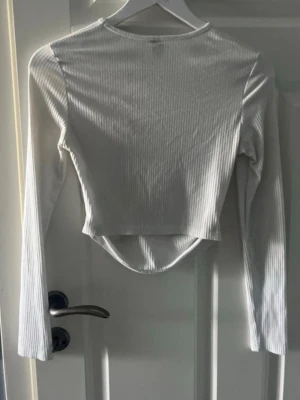 Vit ribbad croppad långärmad topp - Jag säljer en vit ribbad croppad topp med lång ärm och rund hals. Toppen har en snygg, något asymmetrisk nederkant och är i ett stretchigt material som sitter tajt. Perfekt att styla med jeans eller kjol.💕