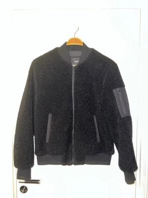 Svart teddyjacka från Pull&Bear - Säljer en svart teddyjacka från Pull&Bear i väldigt fint skick. Jackan är sparsamt använd och har inga synliga skador eller slitningar. Den är både varm och bekväm med stilrena detaljer som dragkedjor och fickor, perfekt för höst och vinter. Finns att hämta i Södertälje.