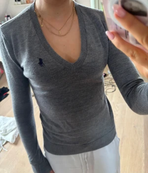 Grå stickad tröja från Ralph Lauren - Snygg grå stickad tröja från Ralph Lauren med djup v-ringning och lång ärm. Tröjan har den klassiska mörkblå loggan broderad på bröstet och ribbade muddar vid ärmslut och nederkant. Perfekt för en stilren och enkel look. Finns ett hål på ena ärmen men det går säkert att sy 