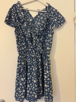 Blå blommig playsuit med volang - Söt blå playsuit med vitt blommönster och volangdetaljer vid halsringningen. Klänningen har korta ärmar, markerad midja och en luftig passform. Perfekt för varma dagar och enkel att styla med sneakers eller sandaler.