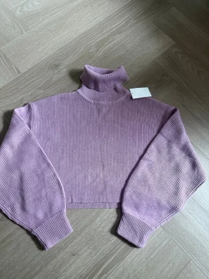 Ljuslila stickad polotröja - Trendig ljuslila stickad polotröja med croppad passform och vida ärmar. Tröjan har ribbstickade muddar och hög polokrage. Perfekt för att skapa en cool och avslappnad look.