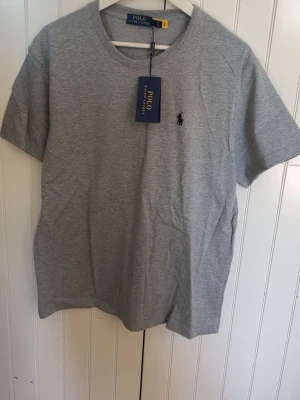 Grå t-shirt från Polo Ralph Lauren - Klassisk grå t-shirt från Polo Ralph Lauren med rund halsringning och ikonisk broderad logga på bröstet. Tillverkad i mjuk bomull och har en normal passform. Perfekt för en stilren och avslappnad look.