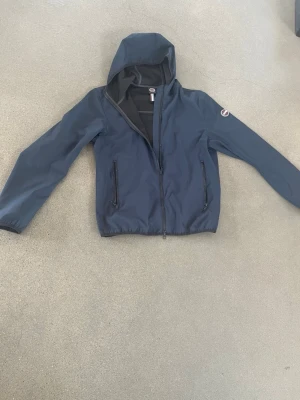 Mörkblå vindjacka med huva - Snygg mörkblå vindjacka från Champion med dragkedja framtill, två fickor med dragkedja och justerbar huva. Jackan har elastiska muddar och logotyp på ärmen samt baktill. Perfekt för blåsiga dagar och enkel att matcha med olika outfits. Använd fåtal gånger