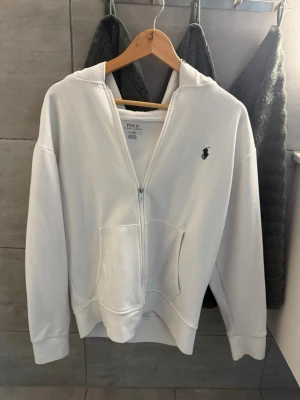 Vit hoodie med dragkedja från Polo Ralph Lauren - Snygg vit hoodie från Polo Ralph Lauren med dragkedja framtill och klassisk huva. Har två stora fickor på magen och den ikoniska loggan broderad på bröstet. Perfekt för en chill och stilren look. Tillverkad i mjukt bomullsmaterial.