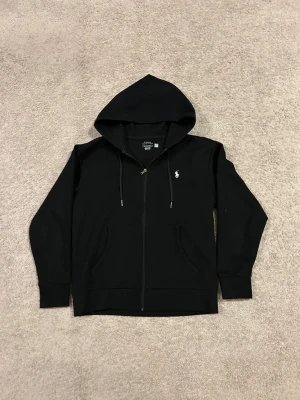 Svart hoodie från Polo Ralph Lauren - Svart hoodie med dragkedja från Polo Ralph Lauren. Klassisk design med huva och dragsko, två fickor framtill och den ikoniska vita logotypen broderad på bröstet. Perfekt för en avslappnad och stilren look.