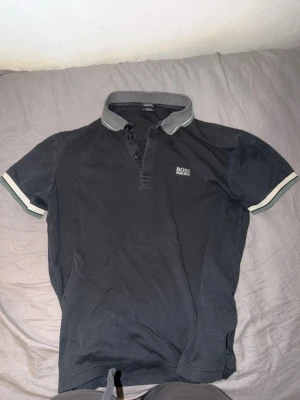 Hugo Boss Piké  - Snygg svart pikétröja från BOSS med grå krage och vita detaljer på ärmsluten. Tröjan har korta ärmar, knappar vid halsen och en klassisk passform. Perfekt för en stilren och avslappnad look.