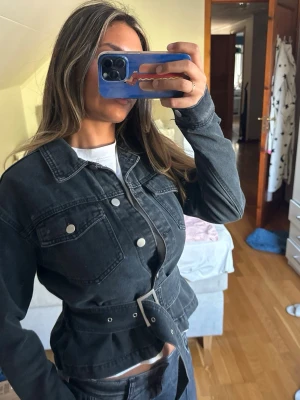 Svart jeansjacka med bälte - Snygg svart jeansjacka med markerad midja och tillhörande bälte. Jackan har två bröstfickor med silverfärgade knappar och klassisk krage. Använt fåtal gånger, kan stylas på olika sätt, även knyta bältet bakom om man önskar 