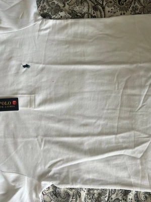 Vit pikétröja från Polo Ralph Lauren - Klassisk vit pikétröja från Polo Ralph Lauren med broderad logga på bröstet och krage med knappar. Tröjan är i bomull och har en stilren och tidlös design som passar till många olika outfits.