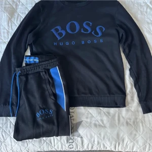 Hugo Boss tracksuit - Snyggt svart loungewear-set från Hugo Boss med blå logga på både tröja och byxor. Tröjan har rund hals och långa ärmar, byxorna har resår i midjan och blå/vita detaljer längs sidan. Perfekt för en avslappnad och stilren look hemma.