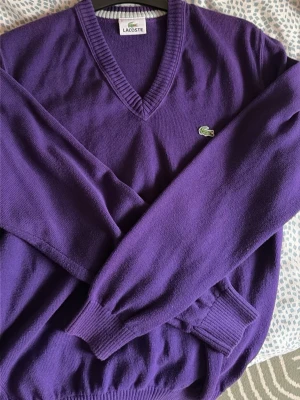 Lila stickad tröja från Lacoste - Snygg lila stickad tröja från Lacoste med V-ringning och klassisk krokodillogga på bröstet. Tröjan har långa ärmar och ribbade muddar vid ärmslut och nederkant. Perfekt för dig som gillar stilrena och färgstarka plagg.