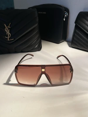 Bruna oversized solglasögon - Snygga oversized solglasögon med bruntonat glas och markerad ram i brunt. Rektangulär form med en cool, modern vibe och gulddetalj vid näsryggen. Perfekta för att lyfta din stil till nästa nivå.