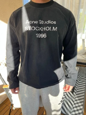 Svart långärmad  Acne Studios L - Svart långärmad topp från Acne Studios med tryck 'Acne Studios STOCKHOLM 1996' på bröstet. Rund halsringning och avslappnad passform. Perfekt för en stilren och enkel look.
