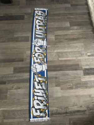 Blåvit halsduk Frihet För Ultras - Snygg halsduk i blått och vitt med texten 'Frihet För Ultras' i stora bokstäver och motiv av knutna nävar. Perfekt accessoar för dig som vill visa ditt stöd på läktaren. Halsduken har fransar i ändarna och coolt streetinspirerat tryck.
