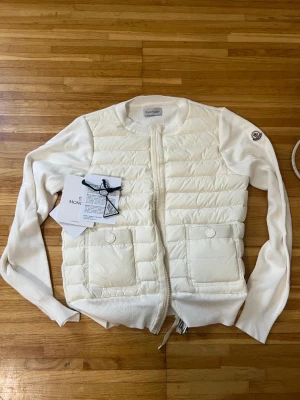 Vit cardiganjacka från Moncler - Snygg vit cardiganjacka från Moncler med quiltad front och stickade ärmar. Jackan har två stora fickor framtill, dragkedja och Moncler-logga på ärmen. Perfekt för lager-på-lager och en clean look. Materialmix av dun och bomull för både värme och stil.