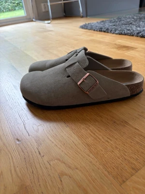 Birkenstock Boston taupe mockatofflor - Säljer ett par Birkenstock Boston tofflor i taupefärgad mocka med justerbart spänne i kopparfärg och klassisk korksula. Perfekta för en chill och stilren look hemma eller ute. Ikonisk design och riktigt sköna att gå i.