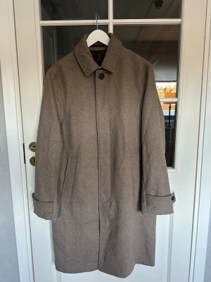 Rock/coat herr - Stilren beige rock med klassisk krage och dold knäppning framtill. Rocken har raka ärmar med knappdetaljer vid ärmslut och två snedställda fickor. Aldrig använd, storlek 50💫