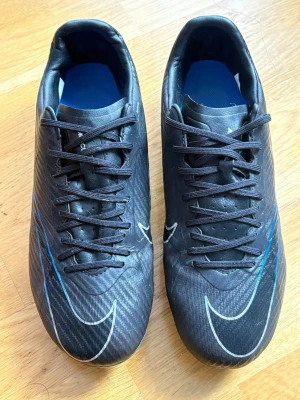 Nike Mercurial svarta fotbollsskor - Säljer ett par Nike Mercurial fotbollsskor i svart med blå detaljer och vita swoosh-loggor. Skorna har snörning, låg profil och mönstrad ovandel i syntetmaterial. Sulan har fasta dobbar för spel på gräs. Perfekta för dig som vill ha snabbhet och kontroll på planen.