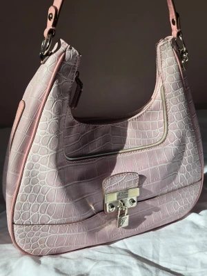 Rosa vintage Guess handväska - Snygg ljusrosa handväska från Guess i krokopräglat skinn med silvriga metalldetaljer och lås framtill från 00-talet. Väskans räm har flagnat något, samt några sömmar har gått upp på framsidan (se bilder)  