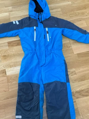 Blå täckbyxa overall Lindberg - Blå och mörkgrå täckbyxa overall från Lindberg med huva, dragkedjor framtill och förstärkta knän. Perfekt för kalla dagar med 10K performance-tyg och praktiska fickor. Snygg och funktionell för vinterns alla äventyr.