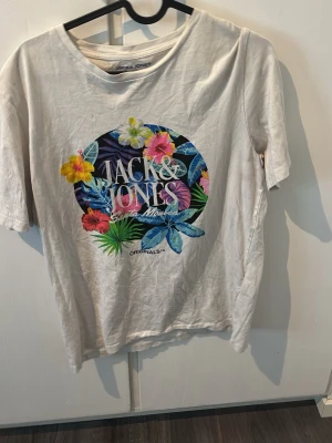 Vit t-shirt Jack & Jones med blommor - Vit t-shirt från Jack & Jones med färgglatt blommönster och tryck framtill. Rund halsringning och korta ärmar. Skön och avslappnad passform, perfekt till sommaren. Materialet är mjuk bomull.