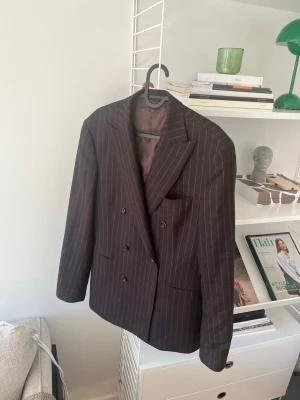 Brun randig dubbelknäppt kavaj - Snygg brun kavaj med tunna vita kritstrecksränder och dubbelknäppning. Klassisk passform med breda slag och tre fickor framtill. Perfekt för en stilren och trendig look.