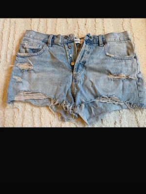  jeansshorts från Zara - Säljer ett par blå jeansshorts från Zara med hög midja och flera slitningar. Shortsen har knappar framtill, klassiska fickor och råa kanter nertill för en trendig look. Perfekta för sommaren och ger en avslappnad vibe. 