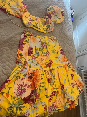 Gult blommigt set med puffärm - Färgglatt tvådelat set med gul botten och stora blommor i lila, orange och vitt. Toppen har puffärmar och resår, kjolen är vid och har volanger. Perfekt för dig som gillar färg och mönster. Materialet känns lätt och luftigt.