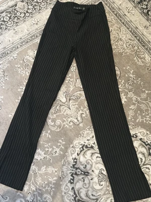 Svarta kostymbyxor med vita ränder - Snygga svarta kostymbyxor  med smala vita kritstrecksränder. Byxorna har rak passform och klassisk midja, perfekta för en stilren look. Materialet känns mjukt och följsamt, vilket gör dem bekväma att bära hela dagen.