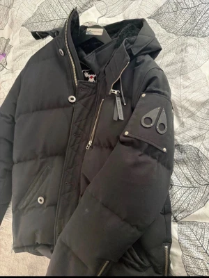 Svart dunjacka från Moose Knuckles - Snygg svart dunjacka från Moose Knuckles med huva, dragkedjor och logotyp på ärmen. Jackan har flera fickor och coola metalldetaljer. Perfekt för kalla vinterdagar och riktigt varm tack vare dunfyllningen.