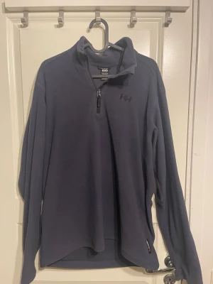 Mörkblå fleece från Helly Hansen - Mörkblå långärmad fleece-tröja från Helly Hansen med halv dragkedja framtill och hög krage. Perfekt för lager-på-lager och outdoor. Diskret HH-logga på bröstet och mjukt material som håller dig varm.