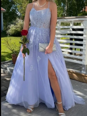 Superfin balklänning  - Säljer en ljuslila balklänning med tunna axelband och öppen rygg (bild 2 & 4). Jag fick så många komplimanger när jag hade den på mig! Klänningen har vackra spetsdetaljer på toppen som ebbar ut på kjolen. Kjolen är i lätt tyg med släp och slits längst ena benet (bild 5). Perfekt för bal, student eller examen! Endast använd en gång. Toppen är korsettliknande med en inbyggd bh (bild 3). 