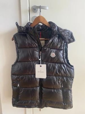 Svart dunväst från Moncler - Snygg svart dunväst från Moncler med glansig finish, huva och dragkedja framtill. Västen har två fickor med dragkedja och Moncler-logga på bröstet. Perfekt för lager-på-lager och streetstyle. 1:1