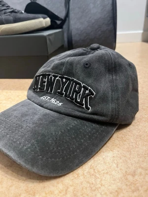 Grå keps med New York-brodyr - Snygg grå keps med böjd skärm och broderad text 'NEW YORK EST. 1625' framtill i svart och vitt. Klassisk modell med justerbar rem bak. Perfekt accessoar för en avslappnad streetwear-look.
