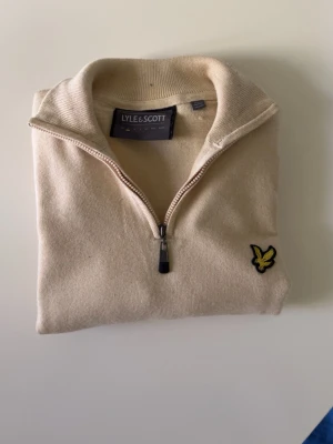Beige halvzip-tröja från Lyle & Scott storlek S - Snygg beige halvzip-tröja från Lyle & Scott med klassisk gul logga på bröstet. Tröjan har hög krage och dragkedja framtill, perfekt för lager-på-lager. Tillverkad i mjukt material som känns skönt mot huden.
