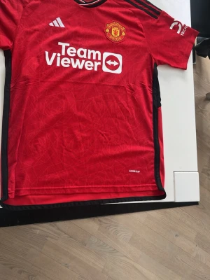 Manchester United matchtröja Adidas - Snygg röd Manchester United matchtröja från Adidas med svarta detaljer och klubbmärke på bröstet. Tillverkad i lätt och ventilerande AEROREADY-material. Tryck med TeamViewer på framsidan och klassiska Adidas-ränder på axlarna.