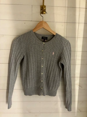 Grå kabelstickad kofta Polo Ralph Lauren - Kabelstickad grå kofta från Polo Ralph Lauren med rund halsringning och knappar framtill. Liten rosa logga broderad på bröstet. Långa ärmar och ribbade muddar. Perfekt för lager på lager och klassisk stil.
