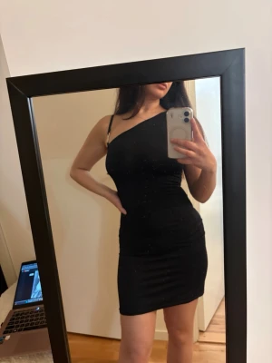 Svart one shoulder klänning  - Snygg svart kortklänning  med one shoulder-design och smal passform. Klänningen har en axelbandslös sida och ett smalt axelband på andra sidan. Perfekt för dig som gillar stilrena och trendiga plagg.