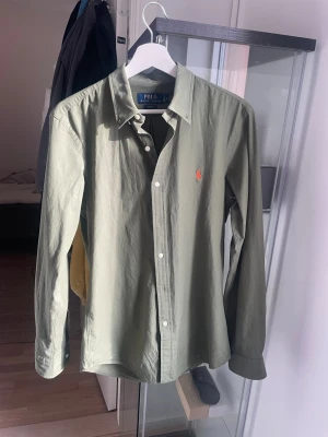 Olivgrön skjorta från Polo Ralph Lauren - Snygg olivgrön skjorta från Polo Ralph Lauren med klassisk krage och knappar framtill. Skjortan har lång ärm och den ikoniska röda logotypen broderad på bröstet. Tillverkad i mjuk bomull för en bekväm känsla. Endast använd en gång säljes pga för liten julklapp! Inga defekter alls utan ser ut som ny, passformen är slim fit så skulle säga att du som är/under 175 men lite mindre i kropps storlek kommer passa super bra i denna!!! Pris går alltid att diskuteras, skriv bara 