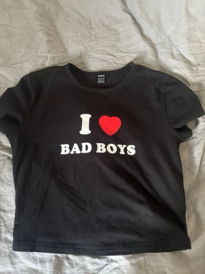 Svart t-shirt med tryck från SHEIN - Svart t-shirt från SHEIN med texten 'I ♥ BAD BOYS' i vitt och rött tryck på bröstet. Klassisk rund halsringning och korta ärmar. Perfekt statement-plagg för dig som vill sticka ut. Pris kan diskuteras, säljer för den är för kort för mig (magtröja typ) 