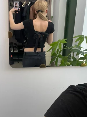 Svart topp med öppen rygg och rosett - Säljer en svart croppad topp med korta ärmar och öppen rygg som knyts med en stor rosett. Toppen har en tight passform och är perfekt för dig som gillar trendiga detaljer. Materialet känns mjukt och stretchigt, troligen bomull eller polyester.