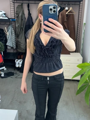 Mörkblå prickig blus med volang - Snygg blå blus med vita prickar och djup v-ringning. Volangdetaljer framtill och smockad midja ger en feminin siluett. Korta ärmar och lätt, luftig känsla – perfekt till jeans eller kjol.