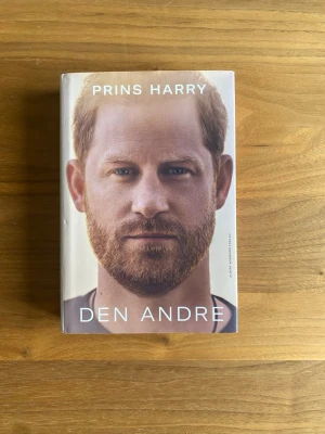 Den Andre av Prins Harry - En inbunden bok med titeln 'Den Andre' av Prins Harry. Omslaget har ett porträttfoto i färg på framsidan och ett svartvitt foto på baksidan. Boken är tryckt på vitt papper och har ett skyddsomslag.